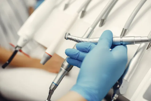 Dentist's gloved hand selects a dental tool from a rack of equipment in a white medical setting.
